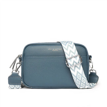 Black leather crossbody bag with a designer geometric strap, featuring teal accents and a rectangular shape, styled as a fashion accessory.