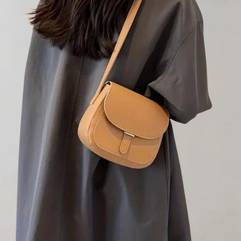 A khaki brown leather crossbody saddle bag with a minimalist design is worn by a woman dressed in a casual outfit featuring a dress shirt and shorts. The bag is positioned near the waist, complementing her attire.