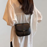 Black leather crossbody saddle bag worn by a woman dressed in a white dress shirt, the bag strap resting on her shoulder and arm.