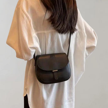 Black leather crossbody saddle bag worn by a woman dressed in a white dress shirt, the bag strap resting on her shoulder and arm.