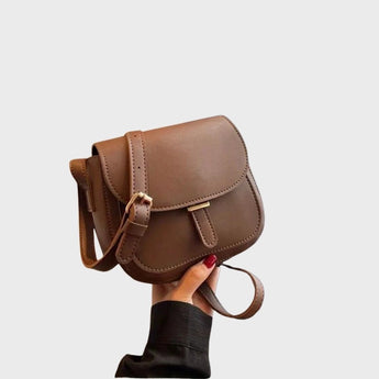 Brown leather crossbody saddle bag with a minimalist design, featuring an adjustable strap and elegant finish, showcased alongside various fashion accessories such as sunglasses and a hat.