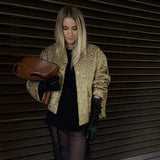 Textured gold metallic bomber jacket worn by a woman, featuring a shiny finish, long sleeves, and a ribbed collar, standing in a pose that highlights the waist and thigh area. 