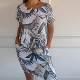 Tropical white botanical print summer shift dress featuring a floral design in white and blue, modeled by a smiling woman. The dress is sleeveless with a round neckline and falls above the knees, suitable for casual summer outings.