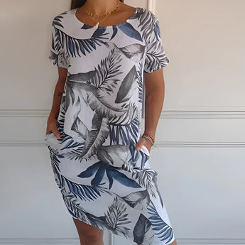 Tropical white botanical print summer shift dress featuring a floral design in white and blue, modeled by a smiling woman. The dress is sleeveless with a round neckline and falls above the knees, suitable for casual summer outings.