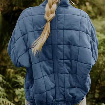 A woman wearing a forest green quilted jacket standing outdoors on grass. The jacket features lightweight insulation with a fitted waist and long sleeves. The woman pairs the jacket with a pair of grey denim jeans.